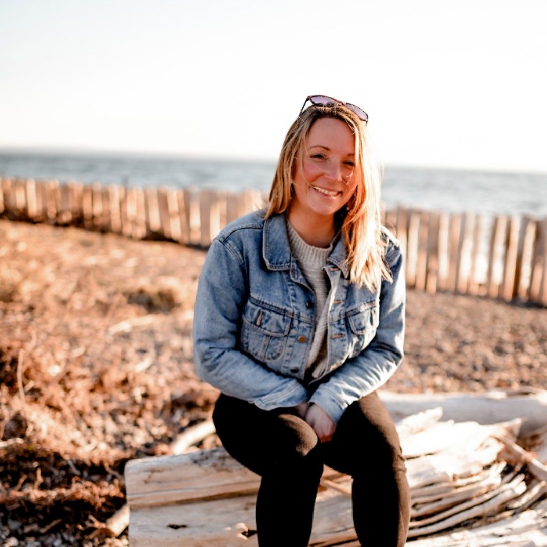 Caroline Landry crédit photo Maude Barriault Sur la plage, une femme est assise sur un bout de bois et souris à la caméra